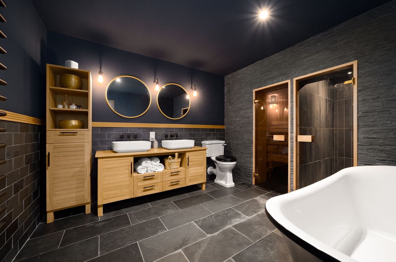 Dark modern bathroom with slate floor tiles, navy blue walls, wooden vanity unit with twin vessel basins, gold-framed round mirrors, freestanding bathtub and walk-in shower room, luxury bathroom photography