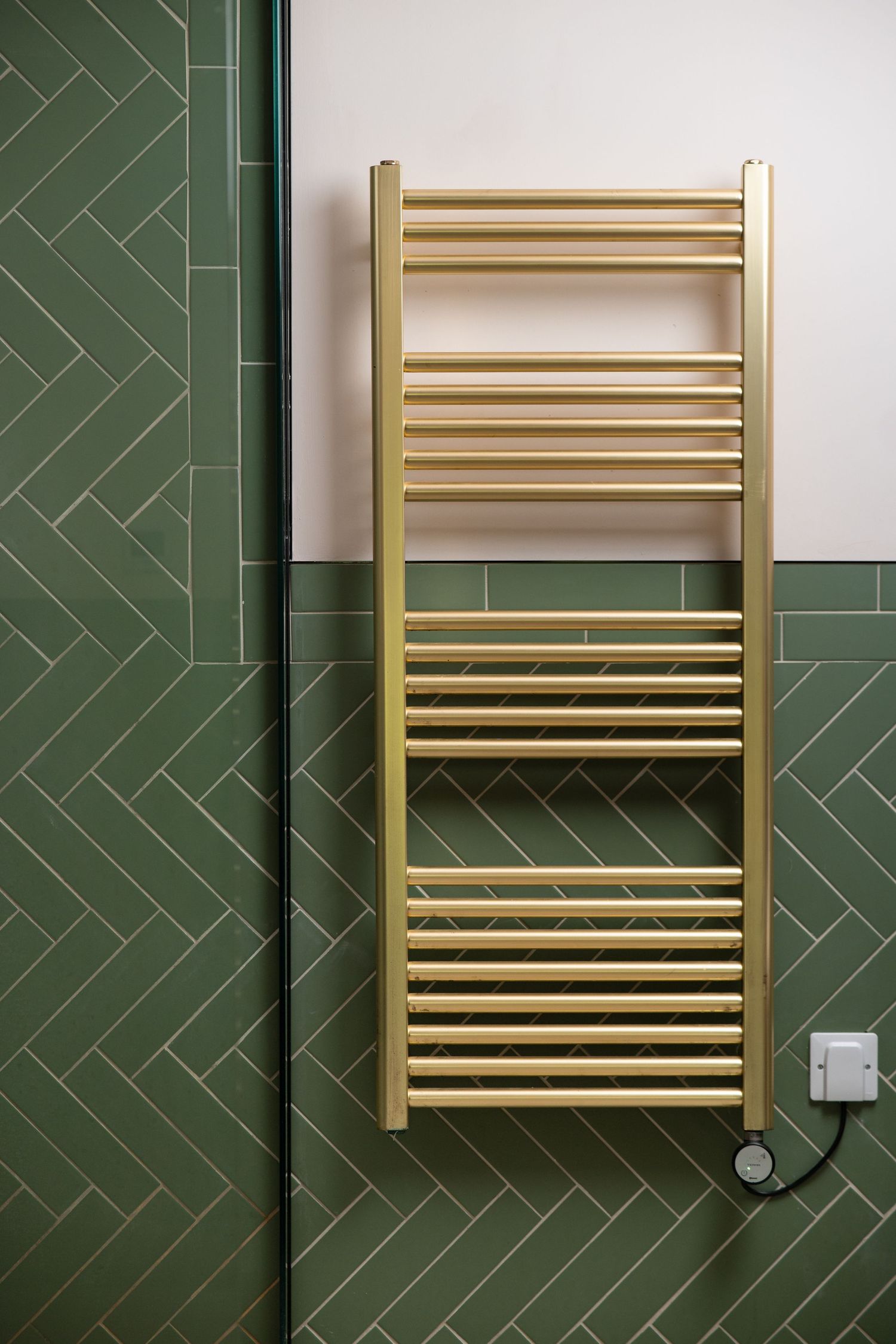 Gold heated towel rail against green herringbone tiles with glass shower screen edge visible, bathroom detail interior photography