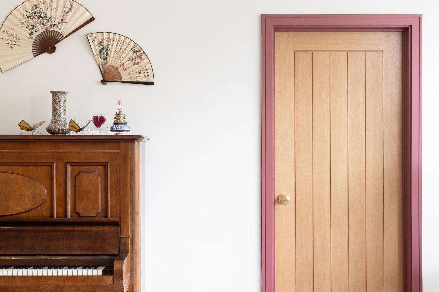 Hallway detail with upright piano, decorative wall fans, ornamental vase and wooden door with pink-painted frame, eclectic interior decor photography