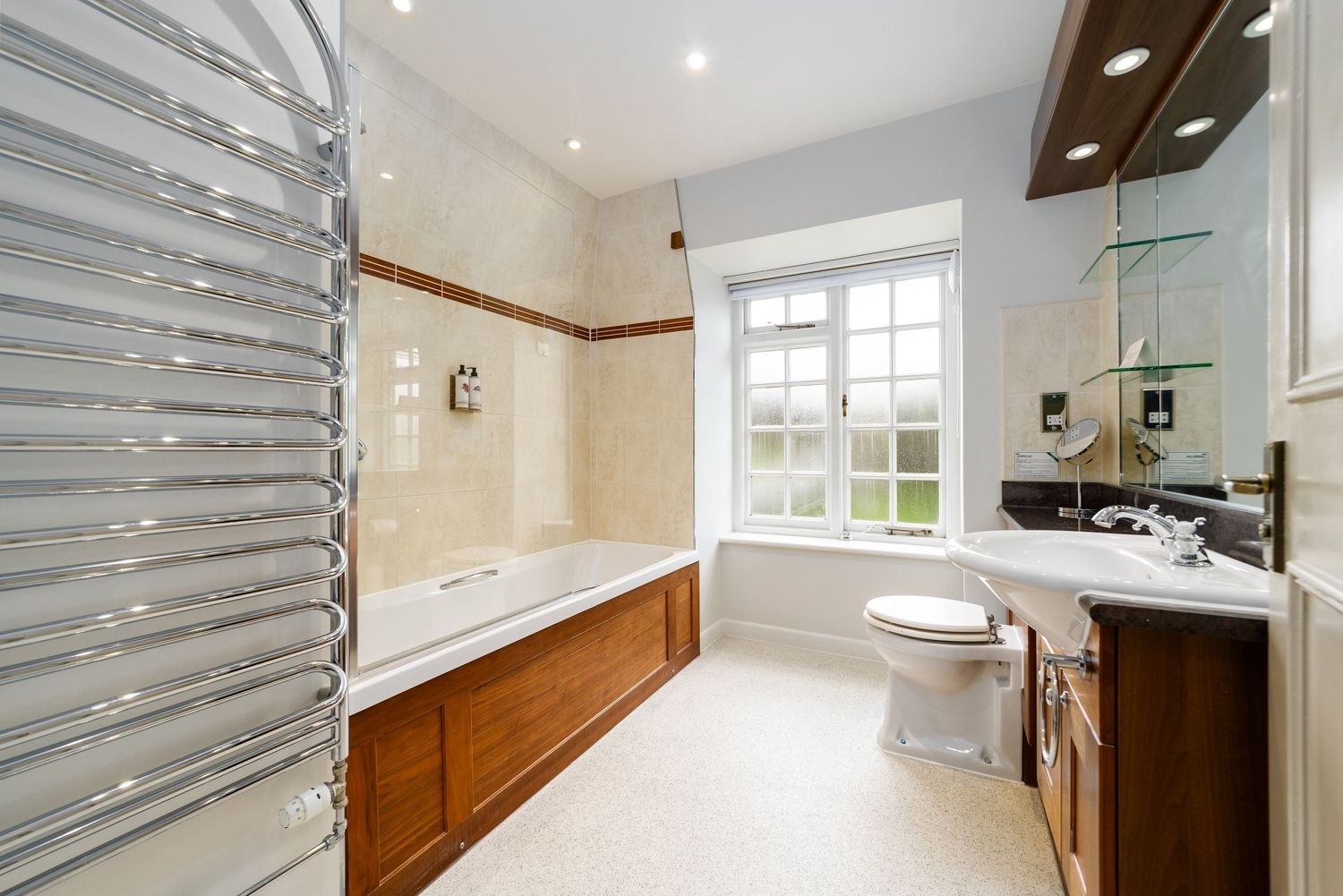 Traditional bathroom with chrome heated towel rail, wooden-panelled bath, beige tiled walls, pedestal basin with dark granite vanity surround and frosted window, bathroom interior photography