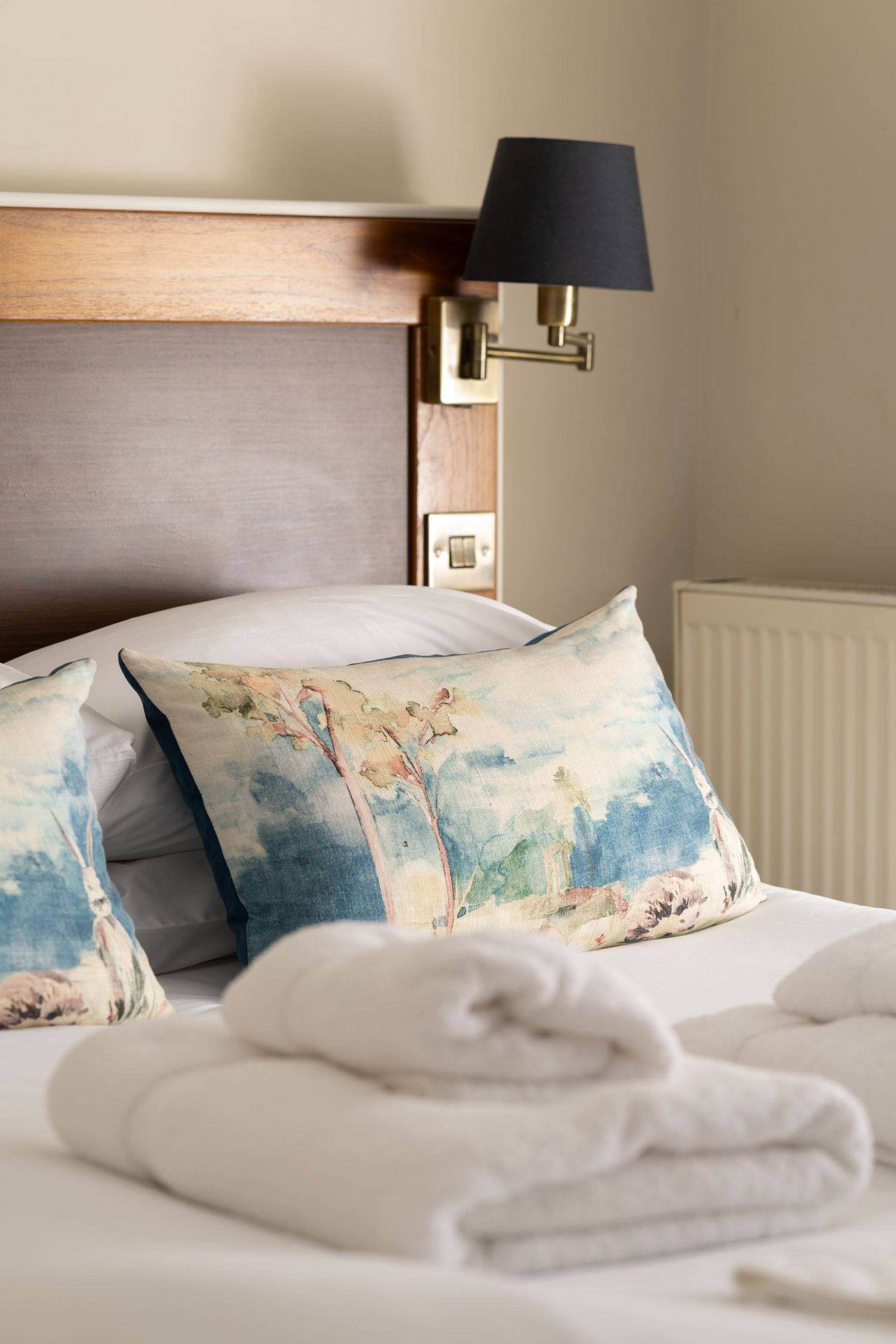 Hotel bed detail with watercolour cushions, folded white towels, wall-mounted reading lamp and wooden headboard, hotel detail photography