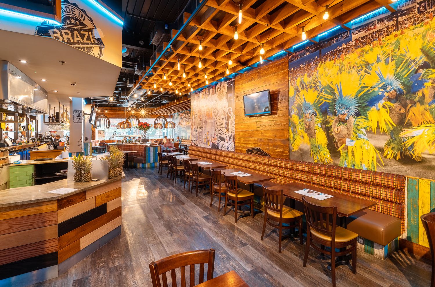 Brazilian restaurant interior with wooden waffle ceiling, vibrant carnival mural, booth seating, rustic wood and blue-tiled bar counter and warm pendant lighting, commercial restaurant photography