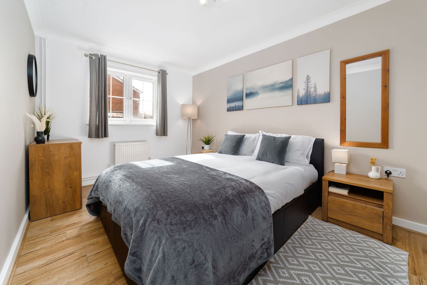 Modern bedroom with grey bedding, oak furniture, landscape triptych artwork, geometric rug and natural daylight from window, real estate interior photography