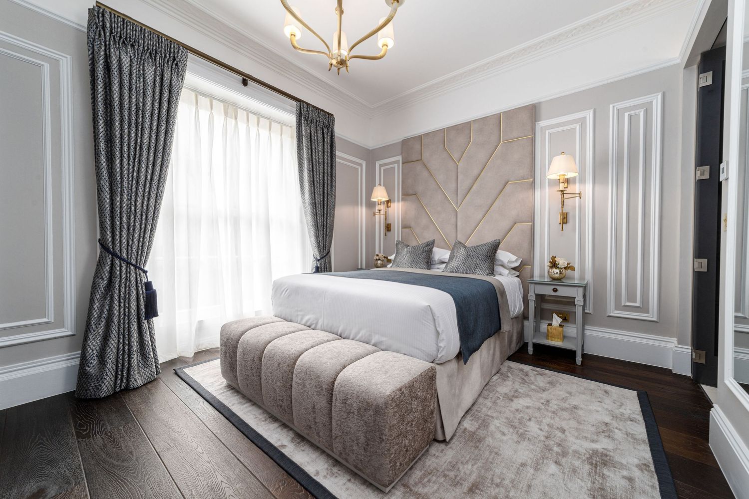 Carlton Hotel luxury bedroom with art deco upholstered headboard, gold chandelier, grey velvet cushions, blue bed runner and ottoman bench on wooden floor, hotel interior photography