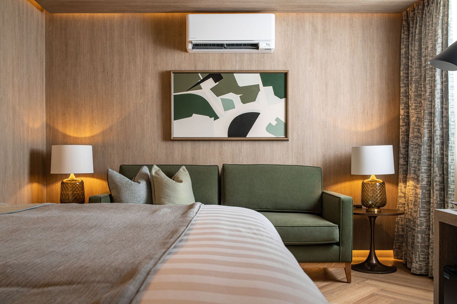 Carlton Hotel bedroom with warm wood-effect wall panelling, green sofa, abstract artwork, amber glass bedside lamps and striped neutral bedding, hotel interior photography
