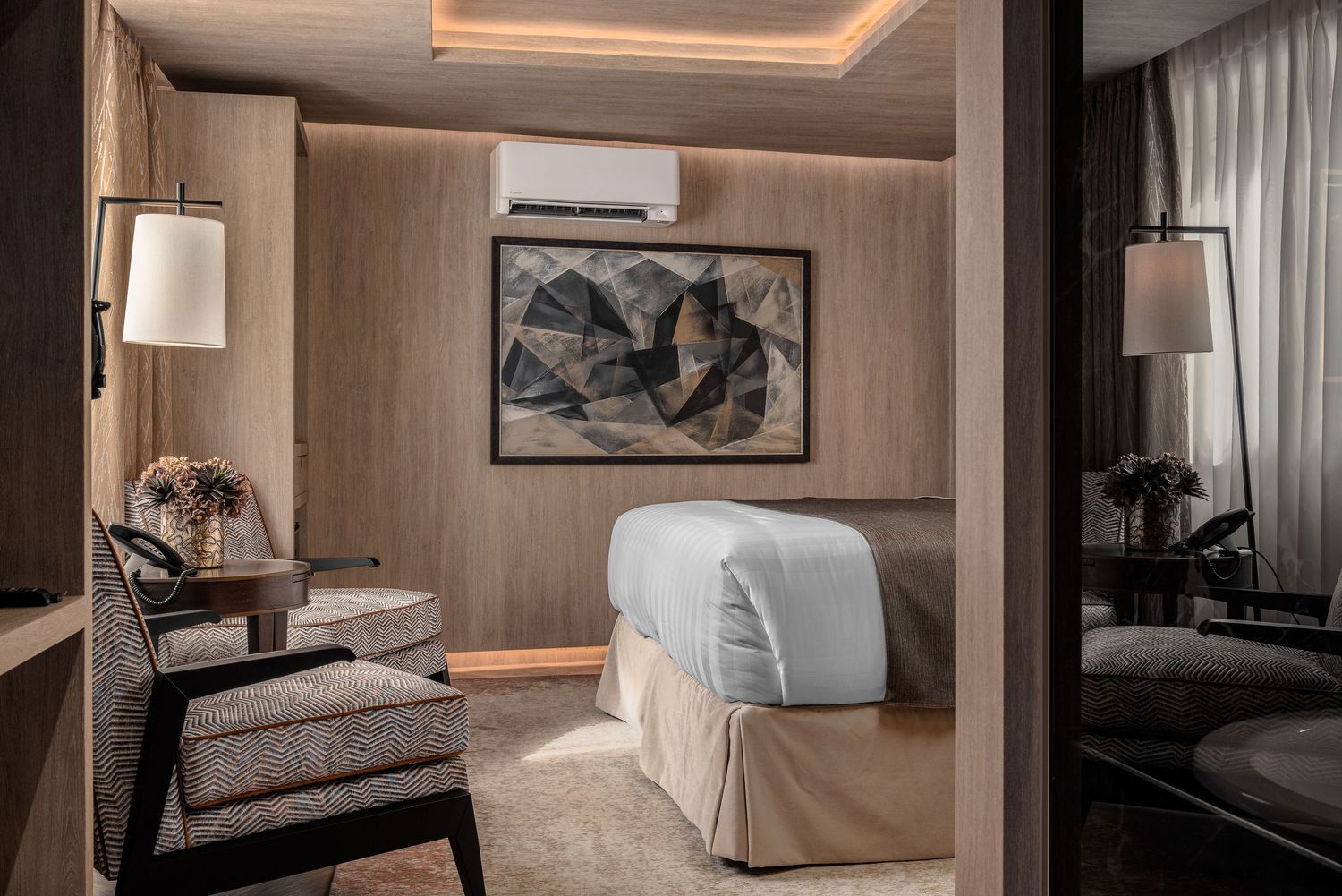 Carlton Hotel bedroom with warm wood panelling, abstract artwork, herringbone upholstered armchair with ottoman, desk area, white bedding and amber glass bedside lamps, hotel interior photography