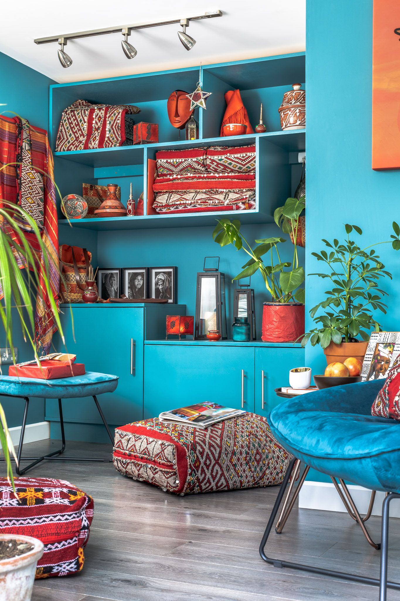 Vibrant teal living room with built-in turquoise shelving, patterned Moroccan-style floor cushions, teal bucket chair and houseplants, eclectic interior photography