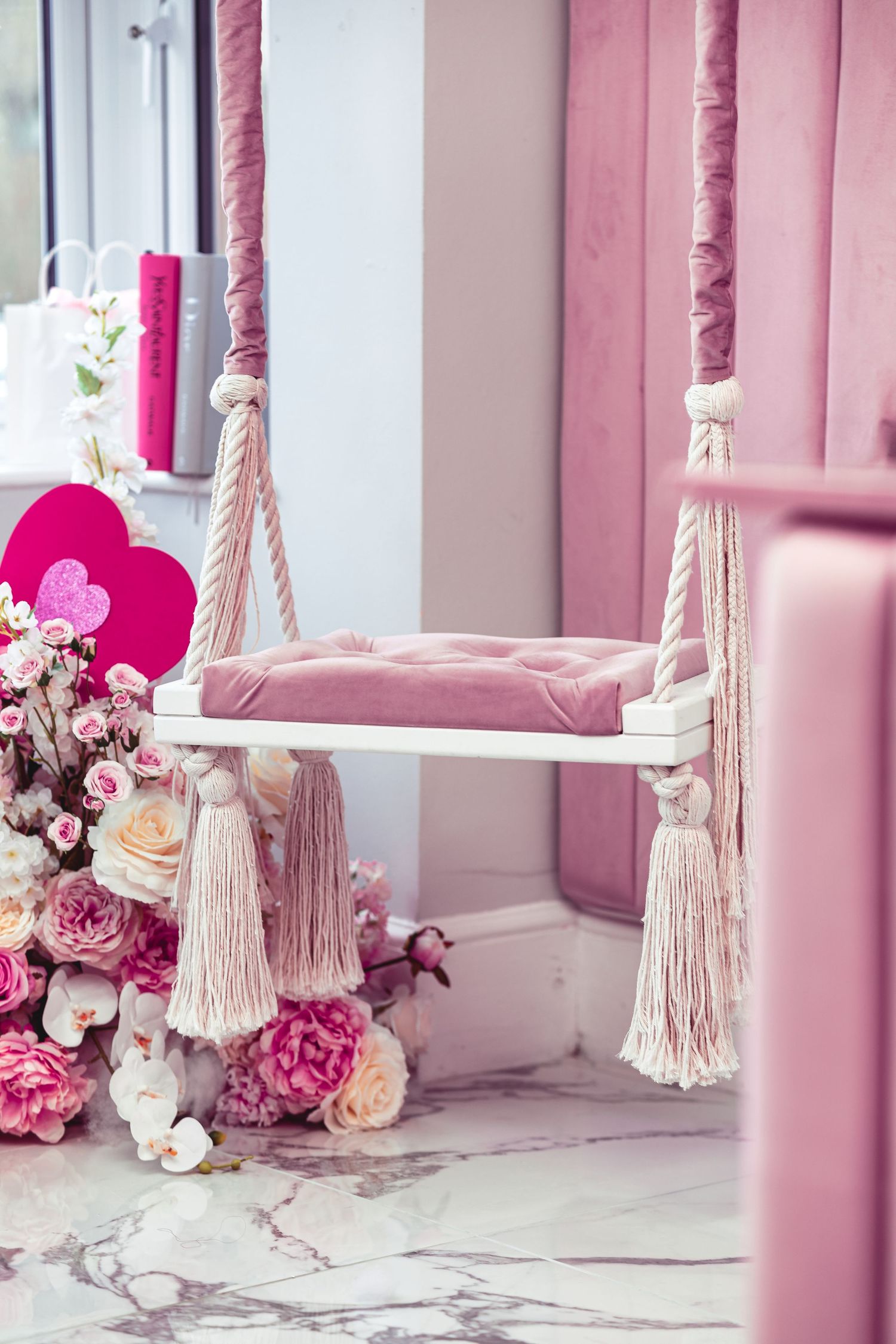 Glam Salon pink velvet rope swing seat with tassel details, surrounded by pink and cream floral arrangements, pink curtains and heart decorations on marble floor, styled salon interior photography