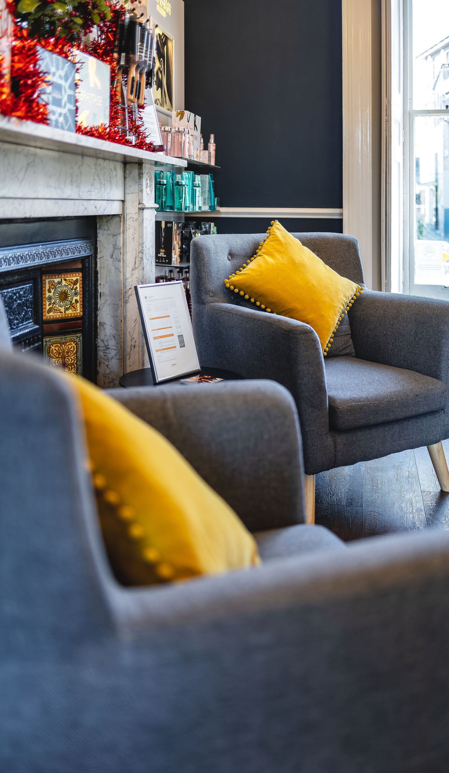 Hair salon waiting area with grey tub chairs, mustard yellow cushions, marble fireplace mantel with hair product display and festive tinsel, commercial salon interior photography