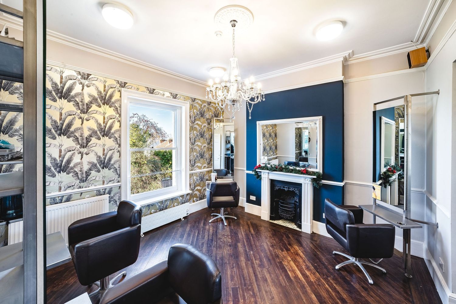 Hair salon interior with black styling chairs, blue feature wall, white fireplace, chandelier, botanical wallpaper and dark wooden floor, commercial salon interior photography