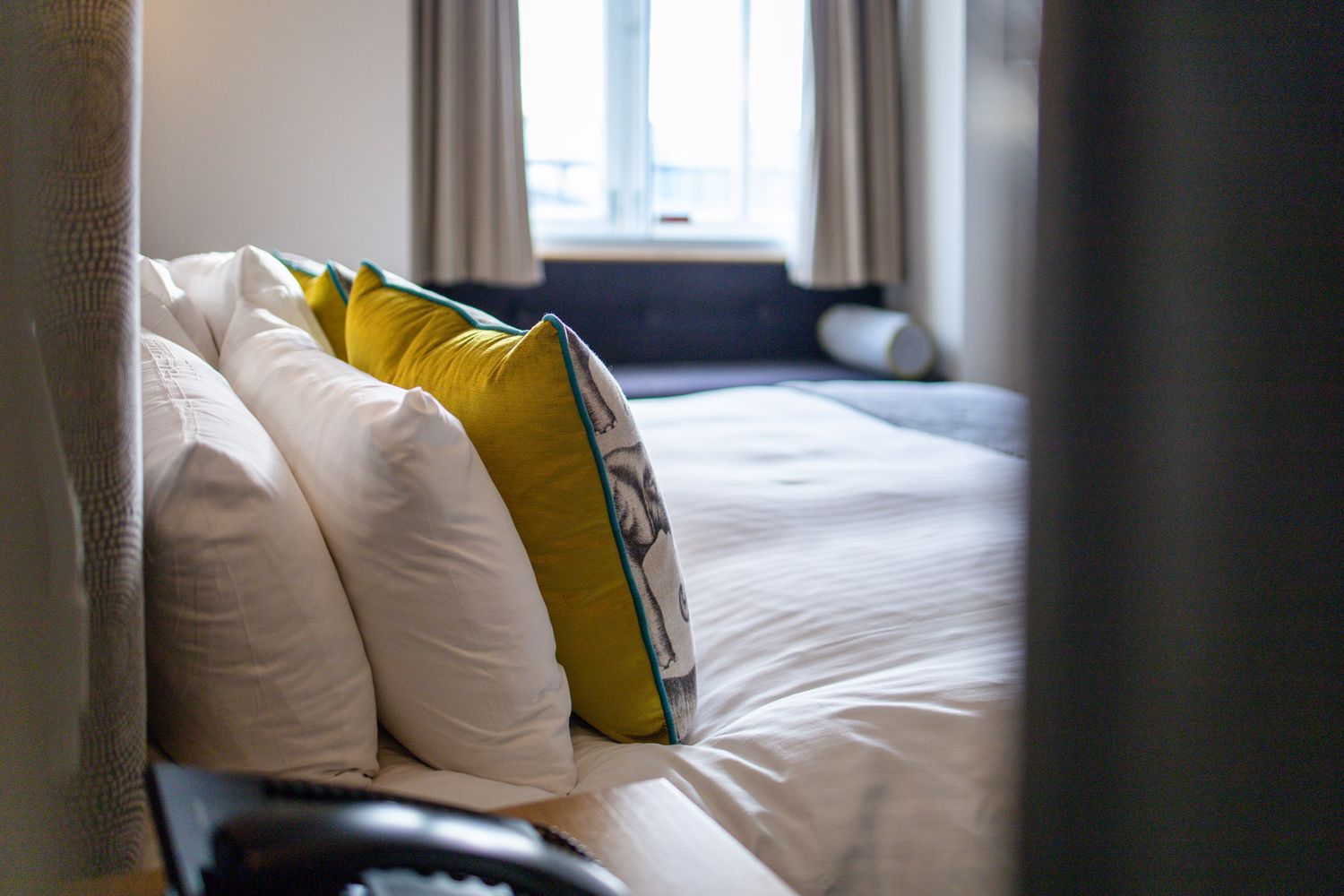Hotel bed detail with white pillows, mustard yellow and illustrated accent cushions, navy sofa beyond and window with curtains, hotel bedroom detail photography