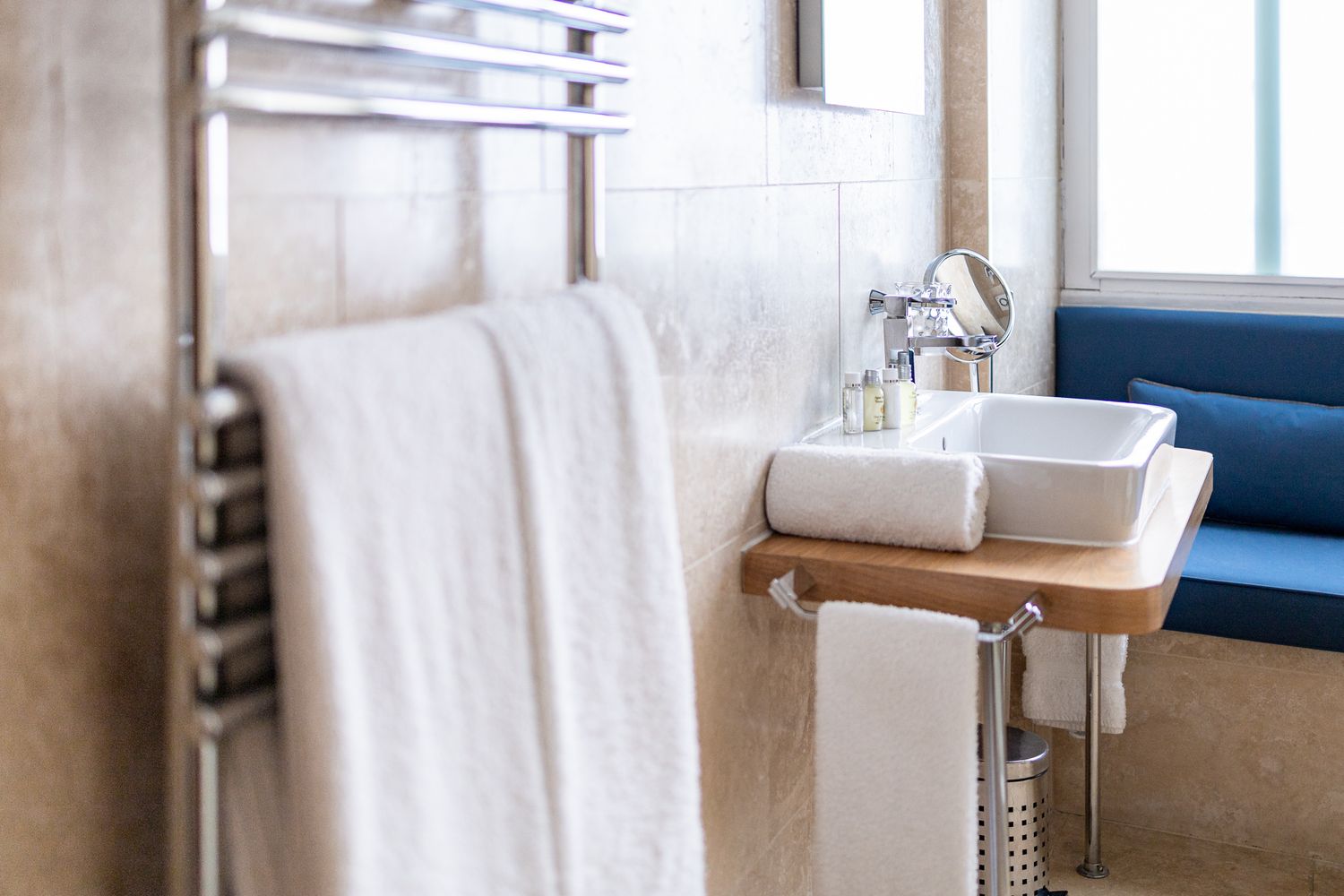 Hotel bathroom detail with chrome heated towel rail, white towels, wall-mounted basin on wooden shelf, toiletries and natural window light, bathroom detail photography