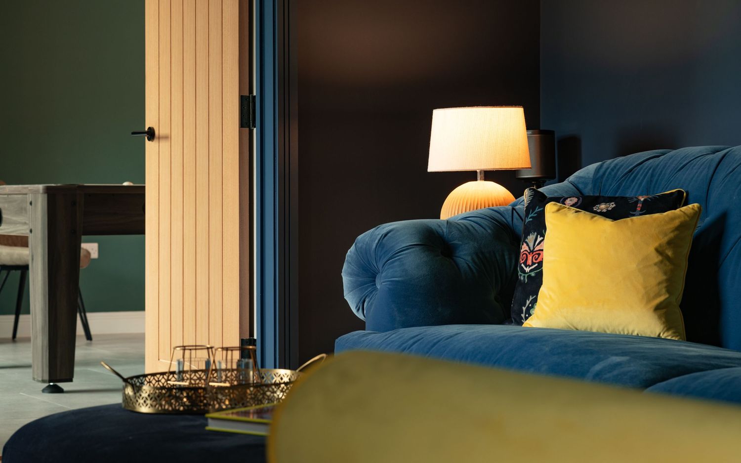 Dark blue living room detail with deep blue velvet Chesterfield sofa, mustard yellow and floral embroidered cushions, table lamp and oak door beyond, moody interior photography