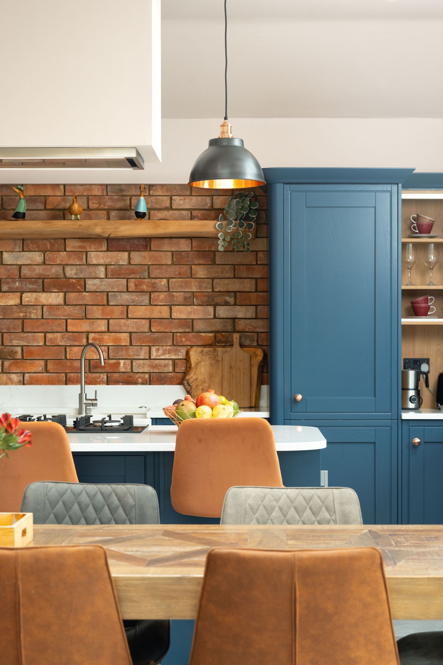 Kitchen-diner with blue cabinets, exposed brick splashback, copper-lined industrial pendant light, tan leather and grey velvet dining chairs and fruit bowl on worktop, kitchen interior photography