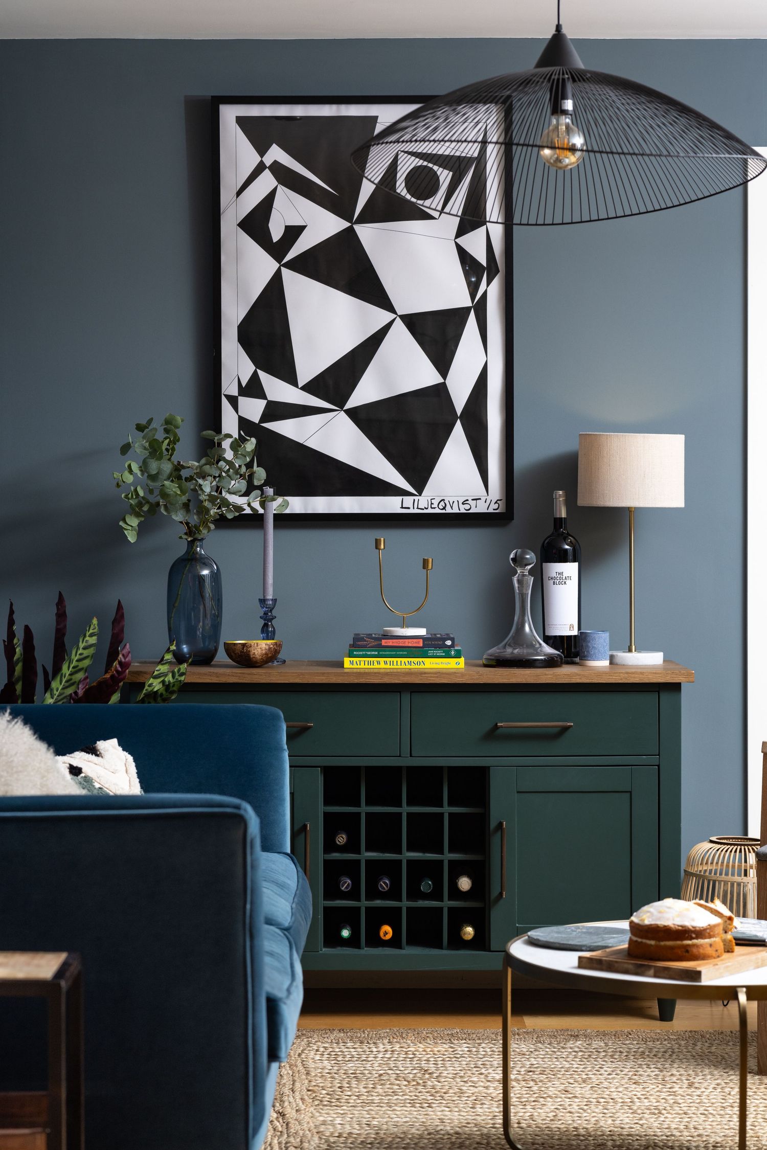 Living room vignette with dark green sideboard and wine rack, black and white abstract artwork, wire pendant light, eucalyptus in a blue glass vase and stacked design books against a blue-grey wall, interior styling photography