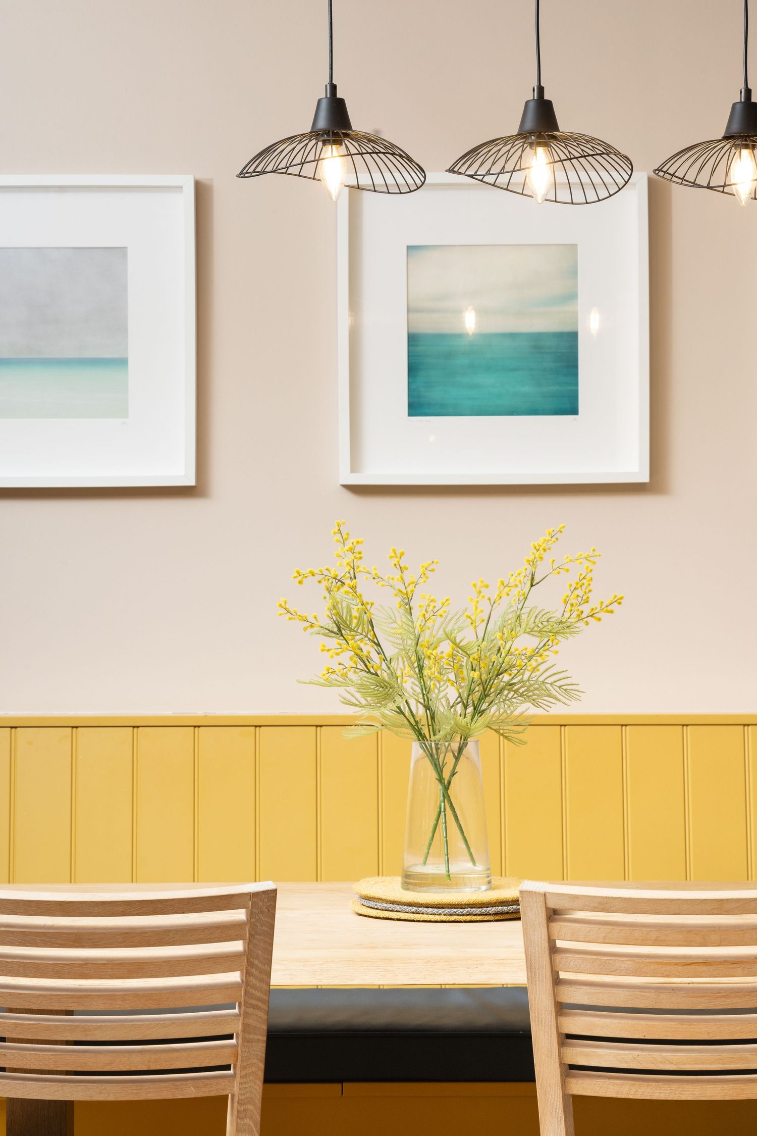 Dining area with yellow panelled dado wall, wooden table and chairs, wire cage pendant lights, coastal seascape artwork and yellow mimosa flowers in a glass vase, interior decor photography