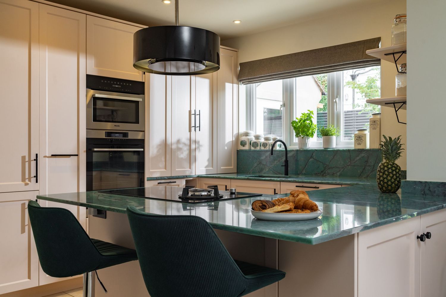 Modern kitchen with cream shaker cabinets, green marble worktops, dark green bar stools, black drum pendant light and built-in ovens, kitchen interior photography