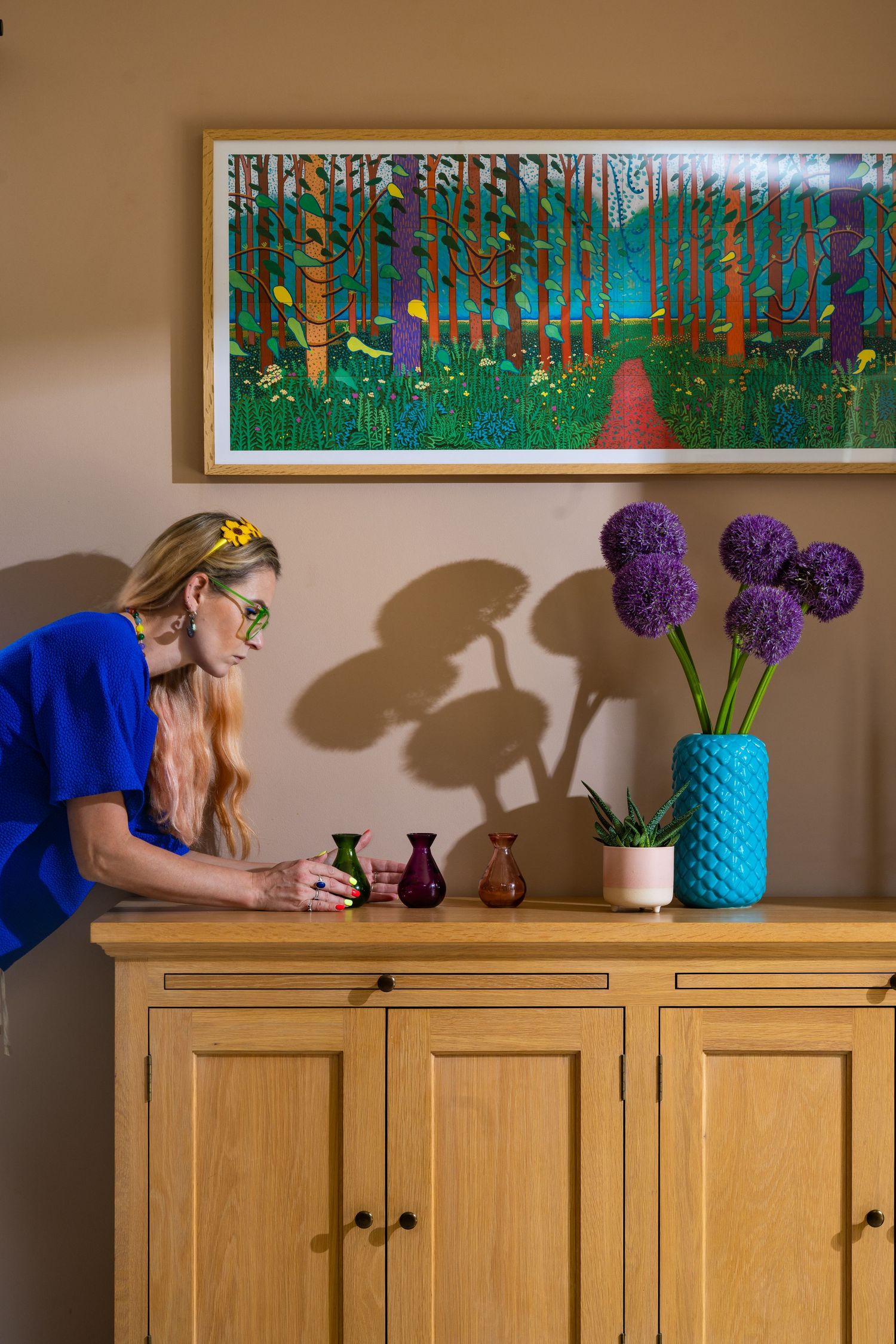 Interior designer placing coloured glass vases on an oak sideboard beneath a large framed woodland print, with alliums in a turquoise vase, interior design photography for Leisha