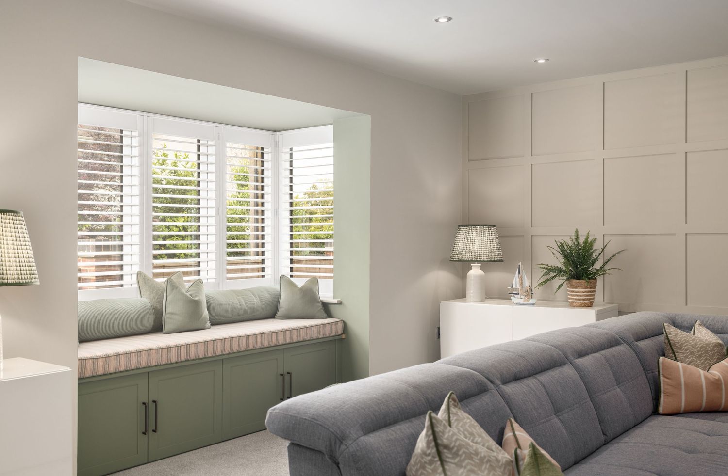Living room with sage green window seat with cushions, grey corner sofa, white panelled wall, plantation shutters and natural light, interior photography