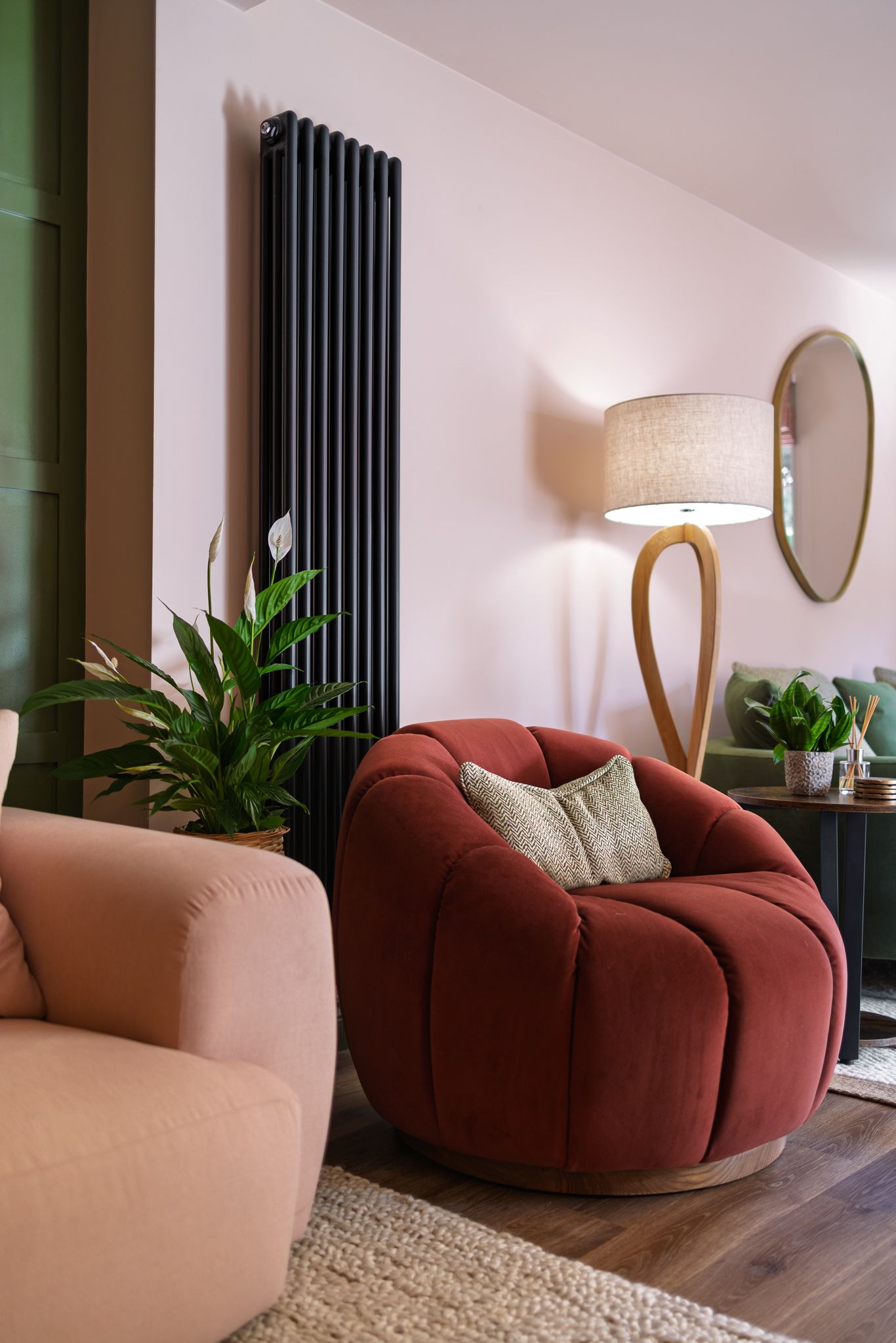 Stylish living room with burgundy tub chair, blush pink sofa, black column radiator, sculptural wooden floor lamp, oval mirror and houseplants, interior design photography
