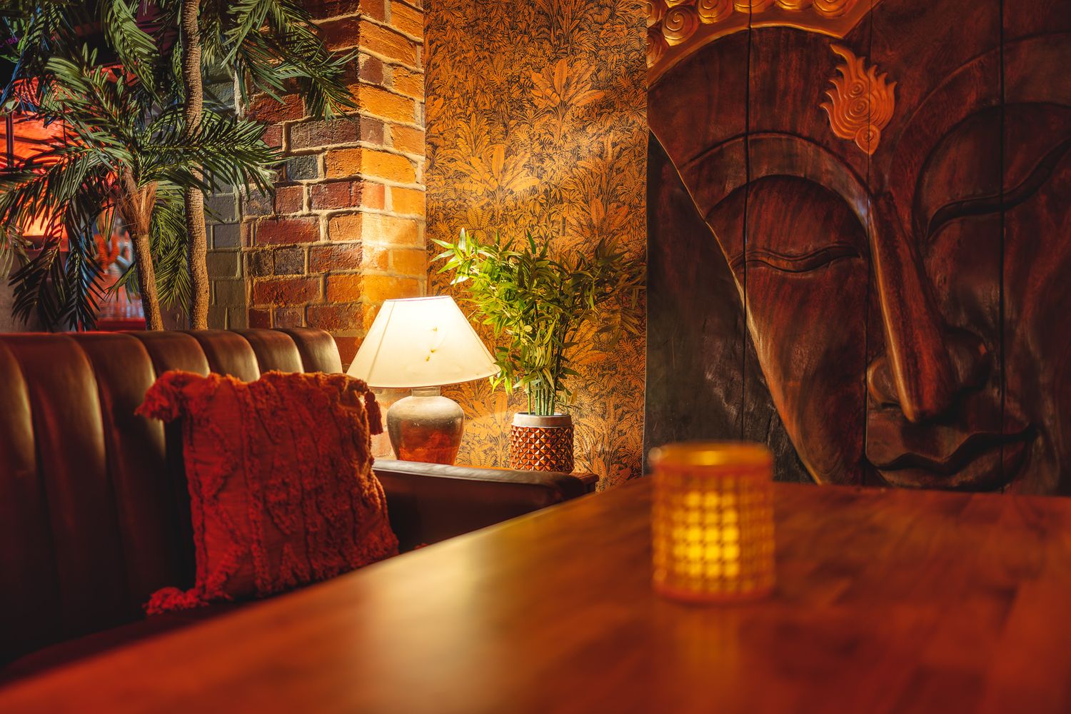 Mango Thai Tapas basement bar booth with leather banquette seating, carved wooden Buddha panel, table lamp, brick wall, red cushion and candlelight, atmospheric restaurant interior photography