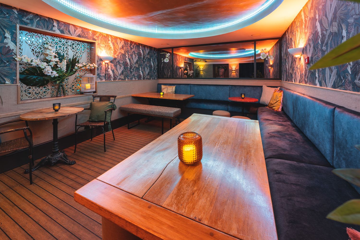 Mango Thai Tapas basement lounge with tropical leaf wallpaper, curved neon ceiling light, teal velvet banquette seating, wooden tables, candles and warm ambient lighting, atmospheric restaurant interior photography