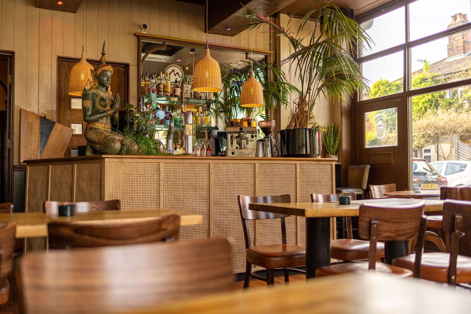 Mango Thai Tapas restaurant bar area with rattan-fronted counter, Buddha statue, wicker pendant lamps, wooden tables and palm plants, commercial restaurant photography