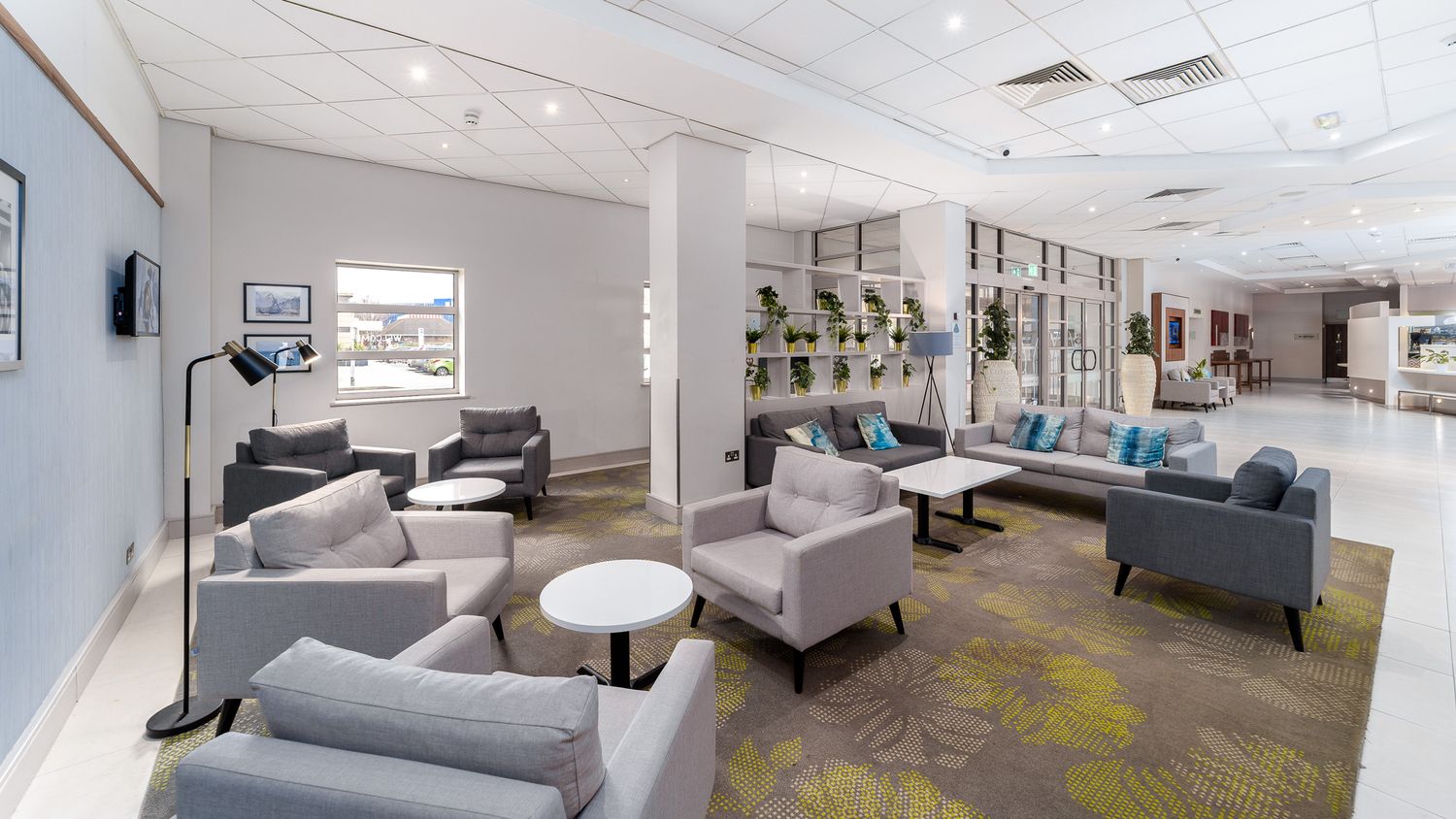 Novotel hotel lobby lounge with grey armchairs and sofas, white coffee tables, patterned carpet, indoor plants on shelving and floor lamps, hotel interior photography