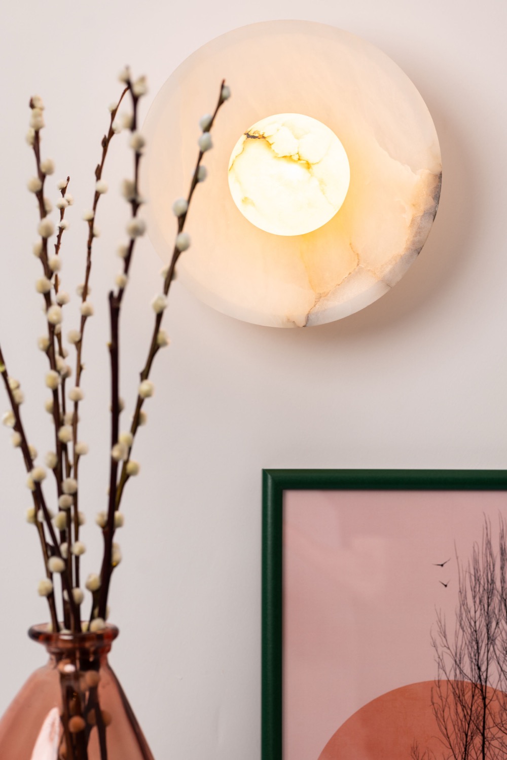 Alabaster disc wall sconce with warm glow above copper vase with pussy willow branches and green-framed pink sunset artwork, interior detail photography