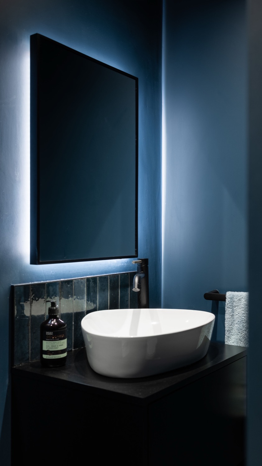 Dark blue cloakroom with LED-backlit rectangular mirror, white vessel basin on dark vanity unit, teal finger tiles, hand wash bottle and folded towel on matt black rail, bathroom interior photography
