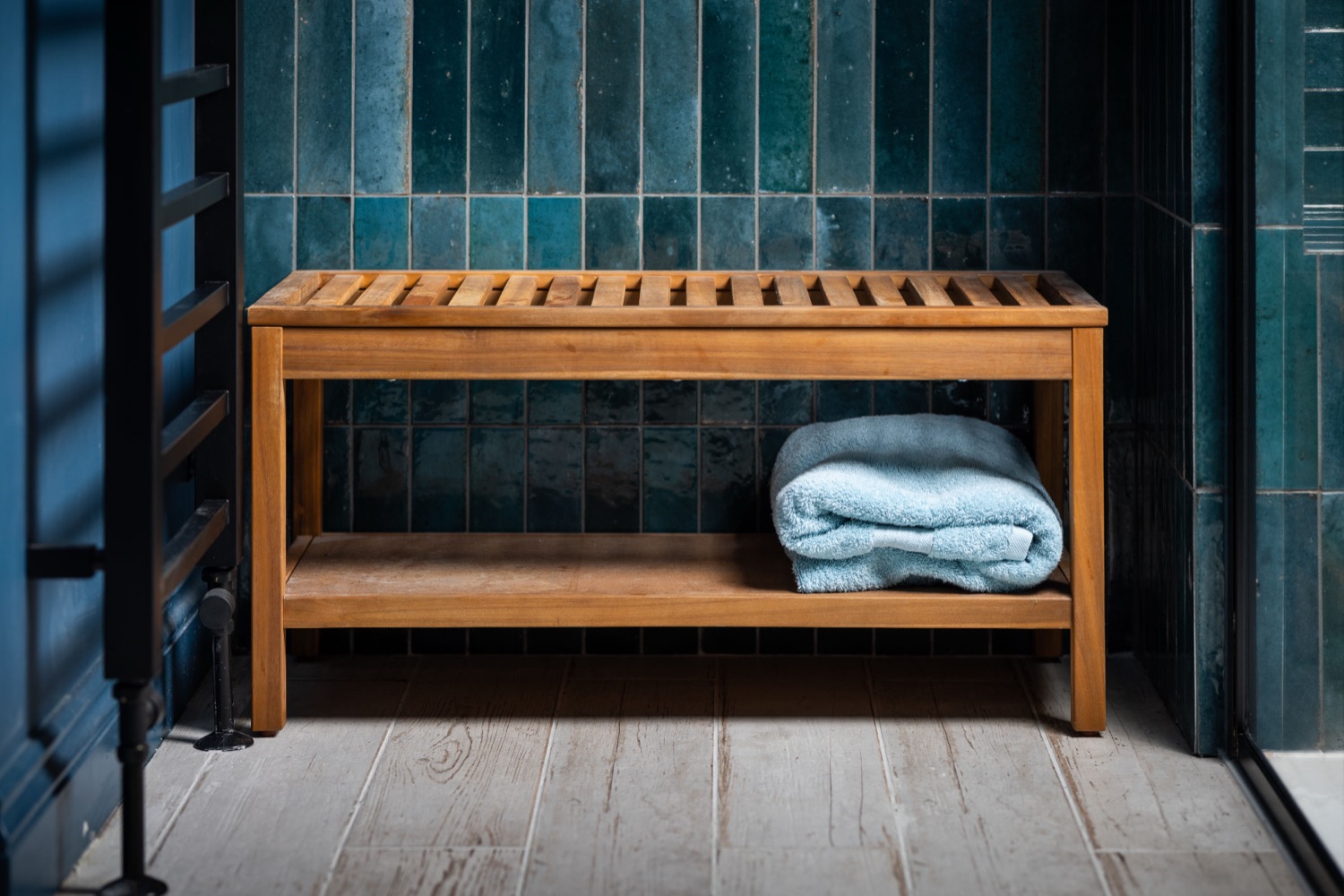 Teak slatted shower bench with folded pale blue towel against teal zellige tiles, black metal shower frame and wood-effect floor, bathroom detail photography