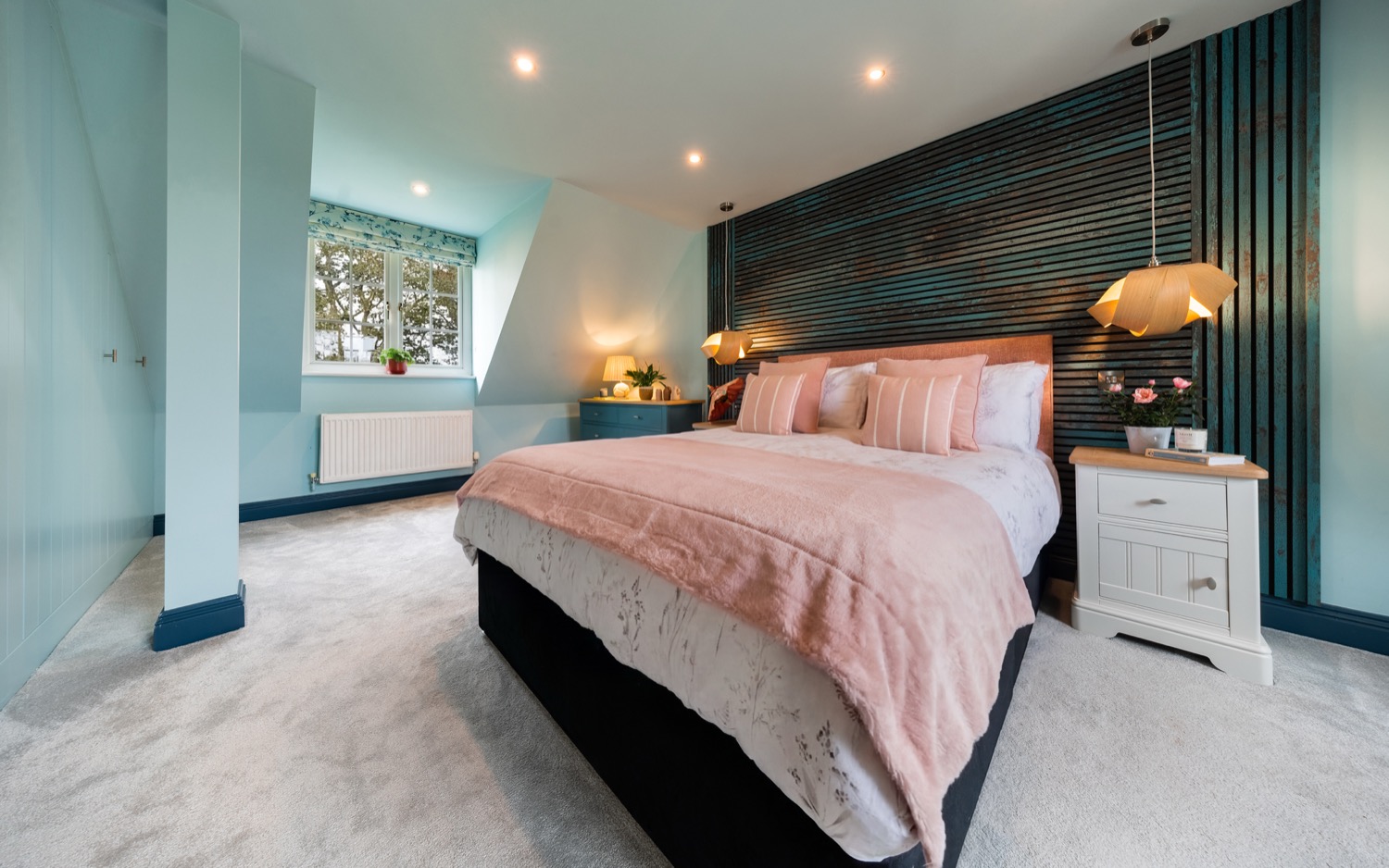 Loft bedroom with dark slatted feature wall, blush pink throw and cushions on king-size bed, duck egg blue walls, dormer window with floral roman blind, geometric pendant lights and white bedside tables, interior design photography