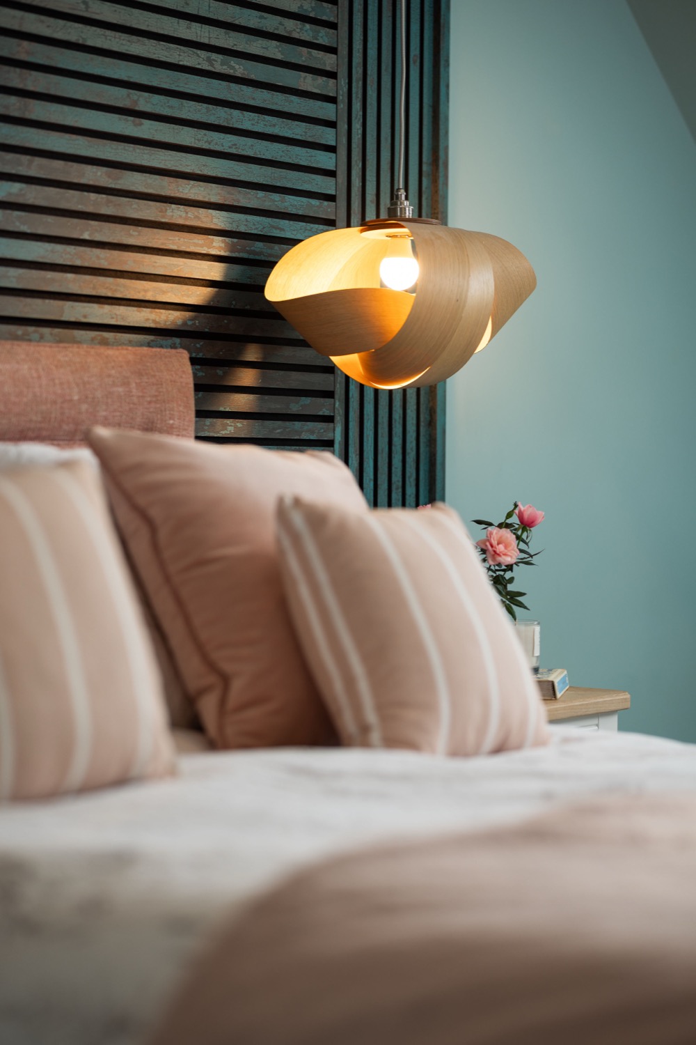 Bedroom detail with sculptural bent-wood pendant light, blush pink striped cushions and bedding, dark slatted headboard panel against teal wall, with pink flowers on bedside table, interior design photography