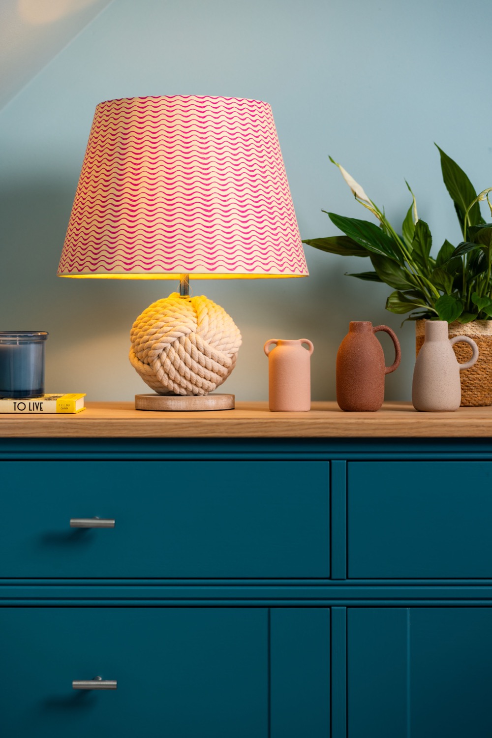 Teal chest of drawers with rope-knot table lamp, pink wave-pattern lampshade, terracotta ceramic vases, peace lily in woven basket and candle against duck egg blue wall, interior styling photography