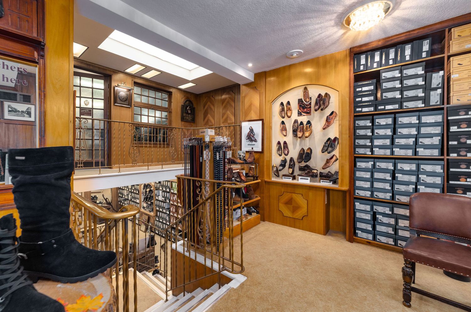 WJ French traditional shoe shop interior with wooden panelling, wall-mounted shoe display, shoe boxes on shelves, brass staircase railing and carpeted upper floor, commercial retail interior photography