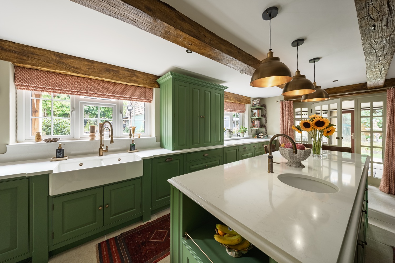 Farmhouse kitchen with sage green painted cabinetry, Belfast sink, brass pendant lights, exposed oak beams and a marble-topped island with sunflowers, designed by Leisha Norman, Sarisbury Green, Hampshire, interior photography