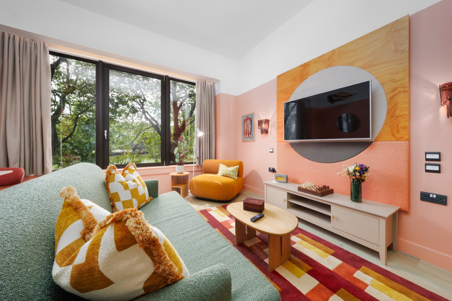 Open-plan room at Second Nature Southampton with coral pink walls, a birch ply television panel, sage green boucle sofa with mustard geometric cushions, burnt orange armchair and a striped wool rug, interior design photography