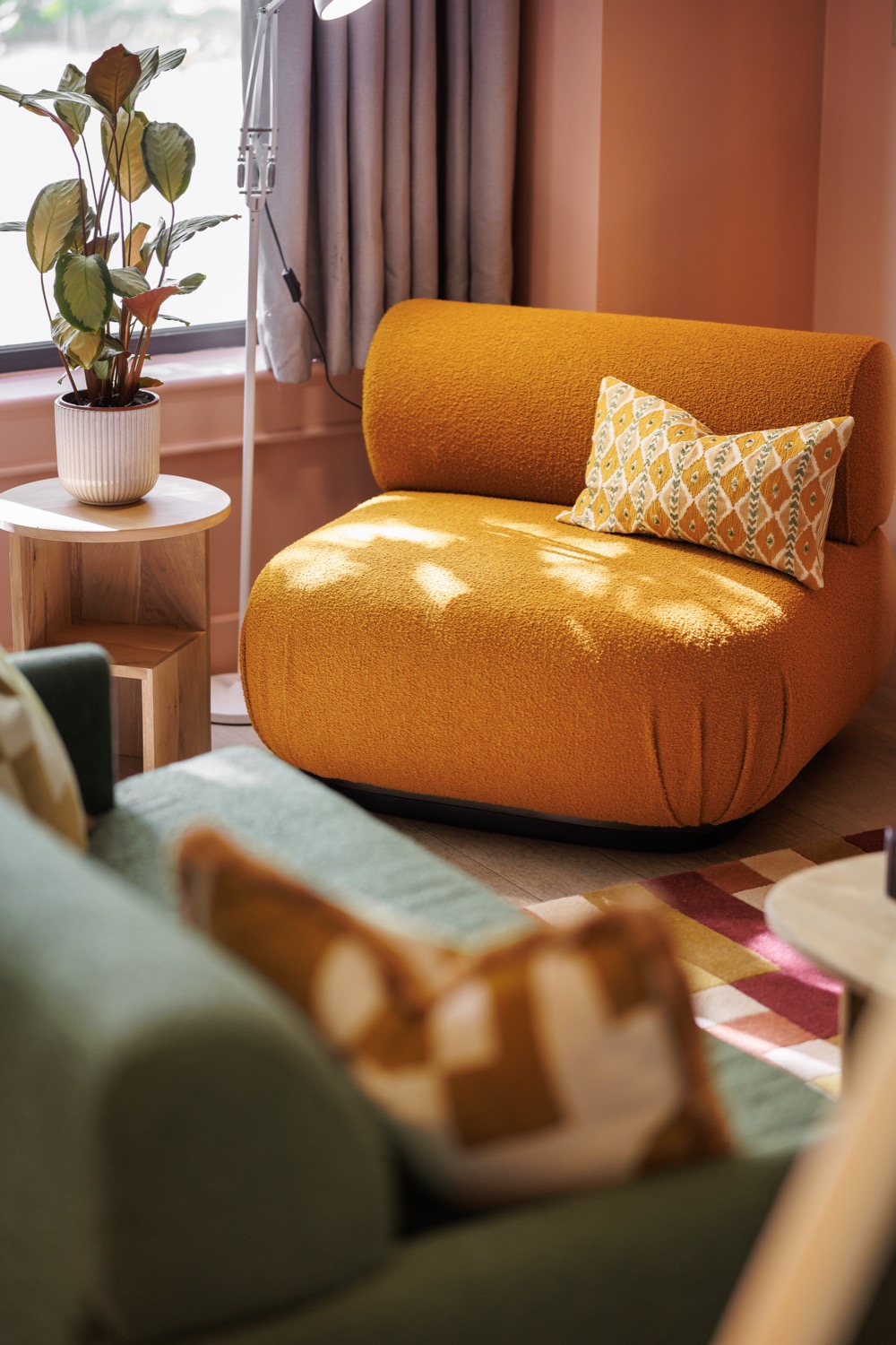 Burnt orange boucle armchair in dappled sunlight with a patterned bolster cushion, a calathea on an oak side table and a sage green sofa in the foreground, Second Nature Southampton, interior design photography