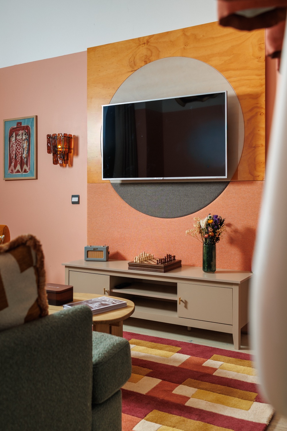 Living area with a wall-mounted TV on a circular plywood and felt panel, coral walls, low sideboard with dried flowers and a chess set, and a geometric rug, Second Nature, Southampton, interior photography
