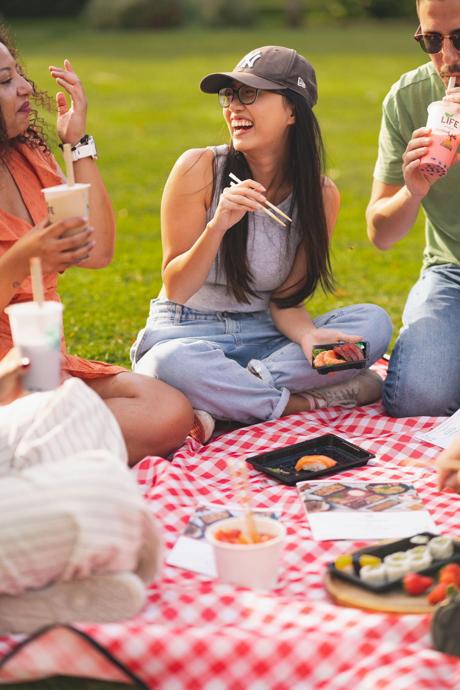 Friends enjoying a sushi picnic on a red gingham blanket in the park for Co-Sushi Southampton, outdoor social lifestyle photography