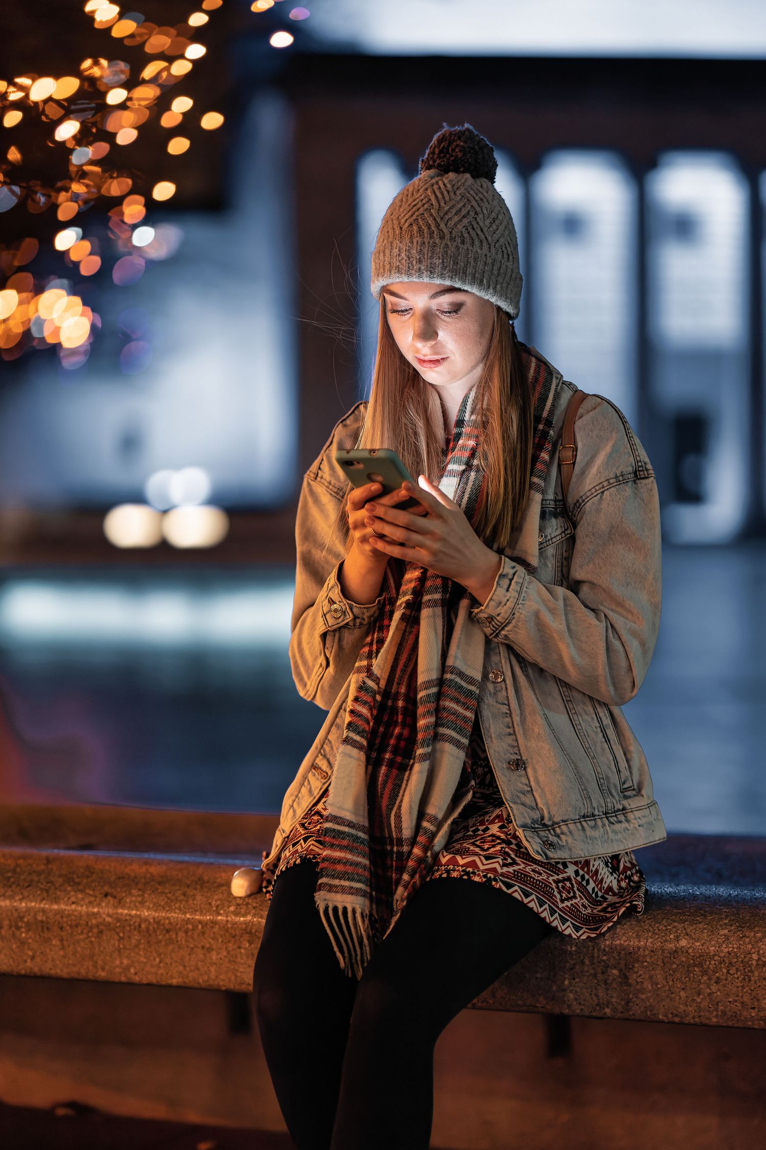 Young woman sitting outdoors at night checking her phone wearing a bobble hat and scarf with fairy lights behind, evening lifestyle photography Southampton