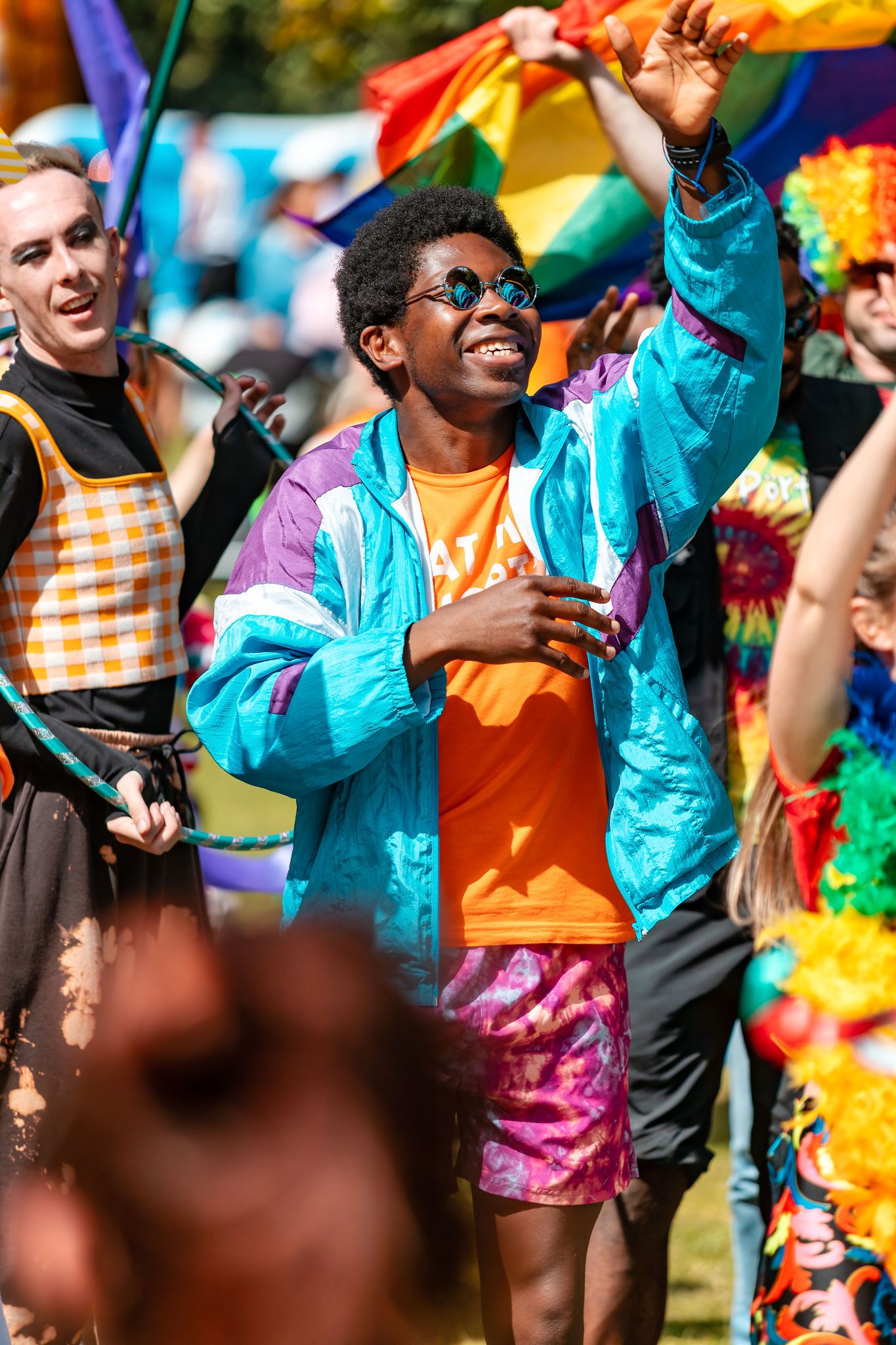Person in a colourful retro windbreaker and sunglasses dancing with rainbow flags at Southampton Pride, candid festival lifestyle photography