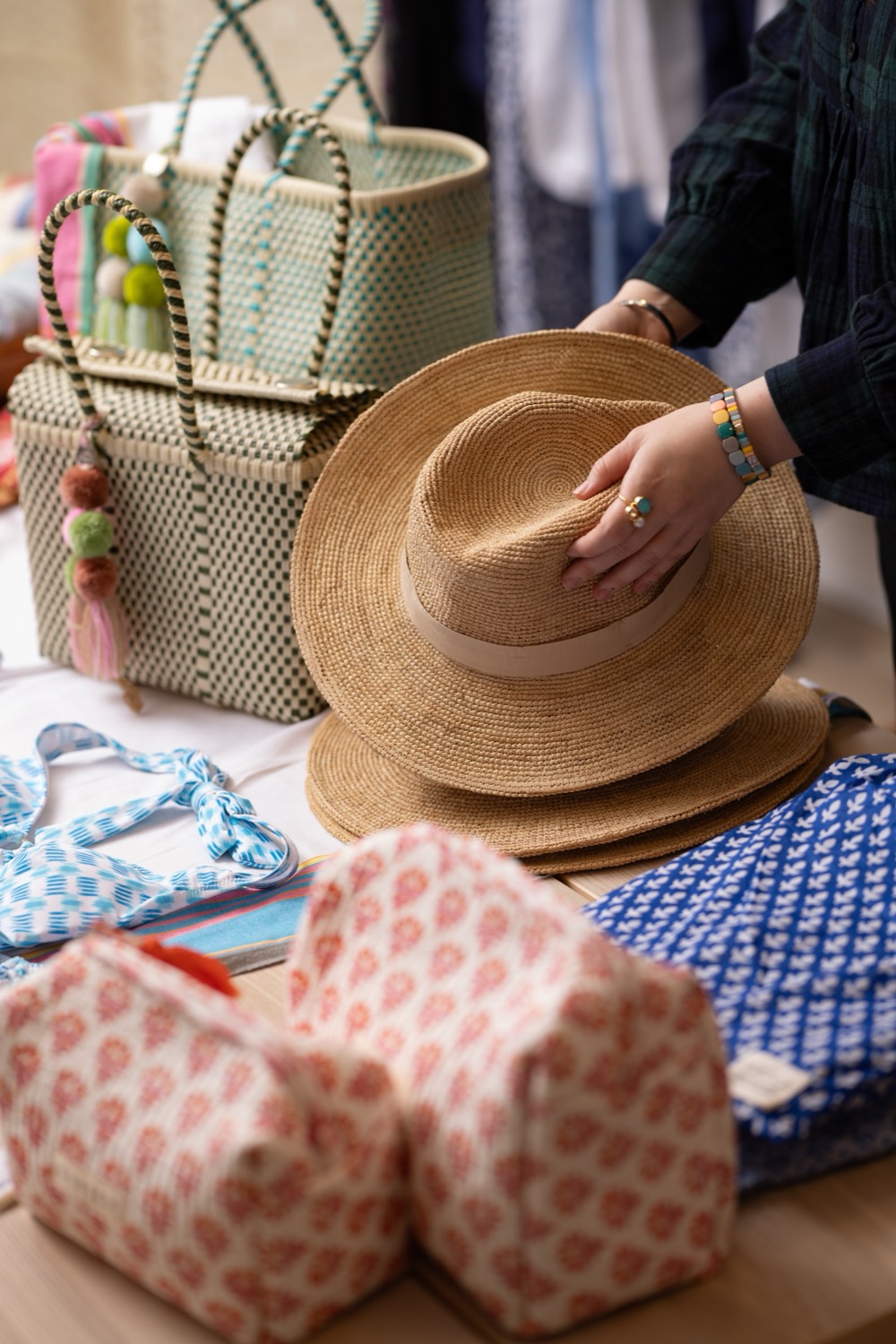 Hands arranging straw fedora hats on a display table with woven tote bags and block-print wash bags at Aspiga Kings Road London, retail lifestyle photography