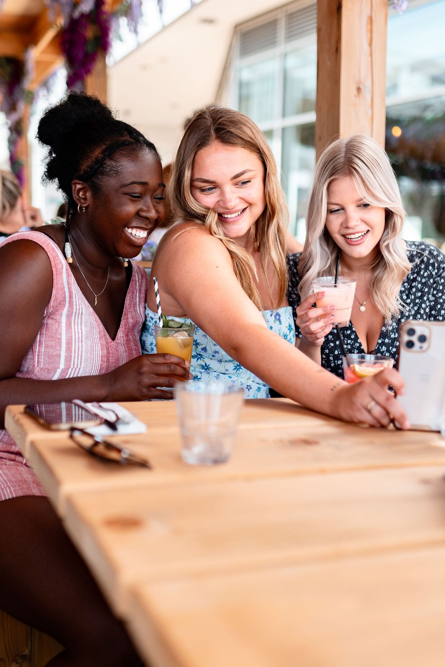 Three friends with cocktails laughing and looking at a phone at an outdoor wooden table, candid social lifestyle photography Southampton