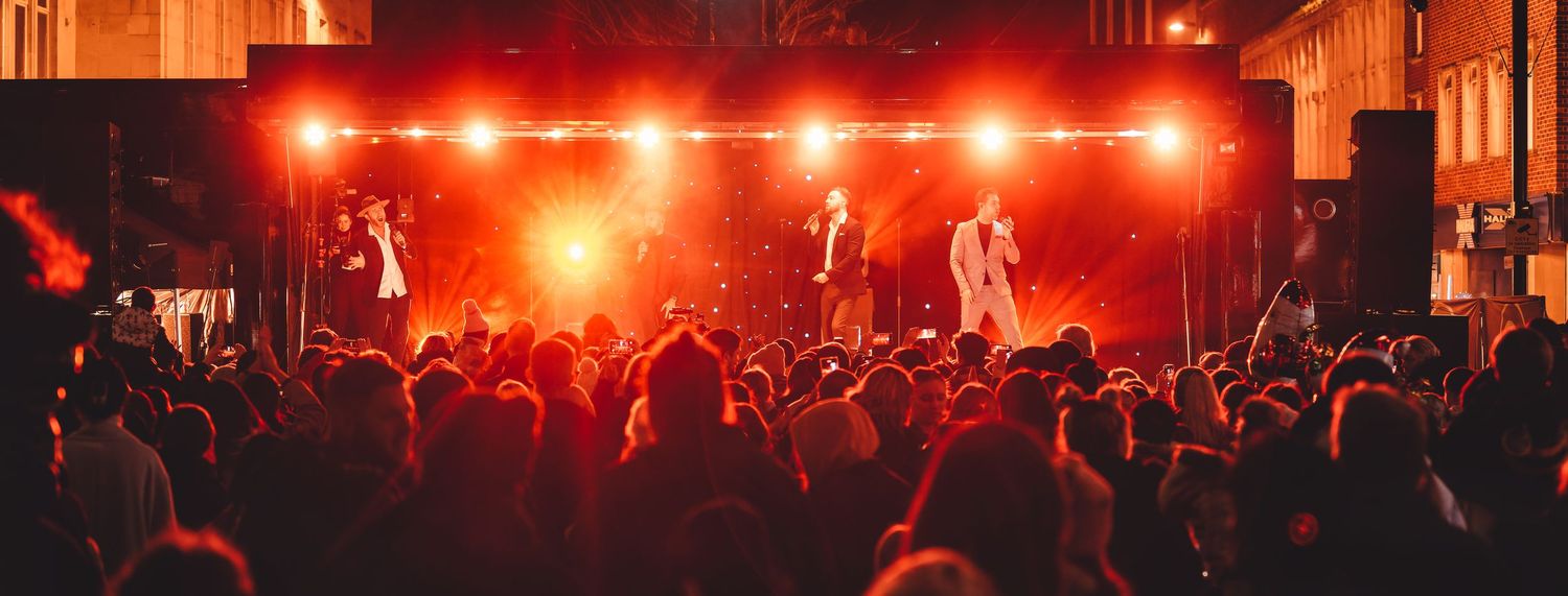 Three suited performers singing on a large outdoor stage with red lighting and a packed crowd at a Countdown to Christmas event, live event photography Southampton