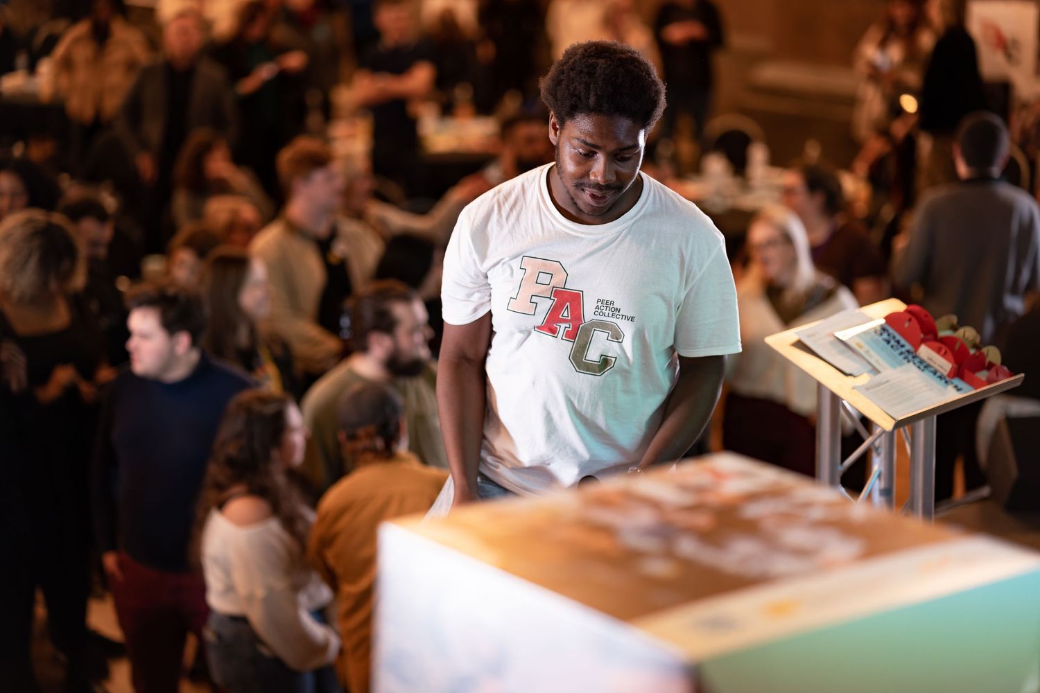 Young man browsing a stall at a busy indoor community event with people socialising in the background, candid event photography Southampton