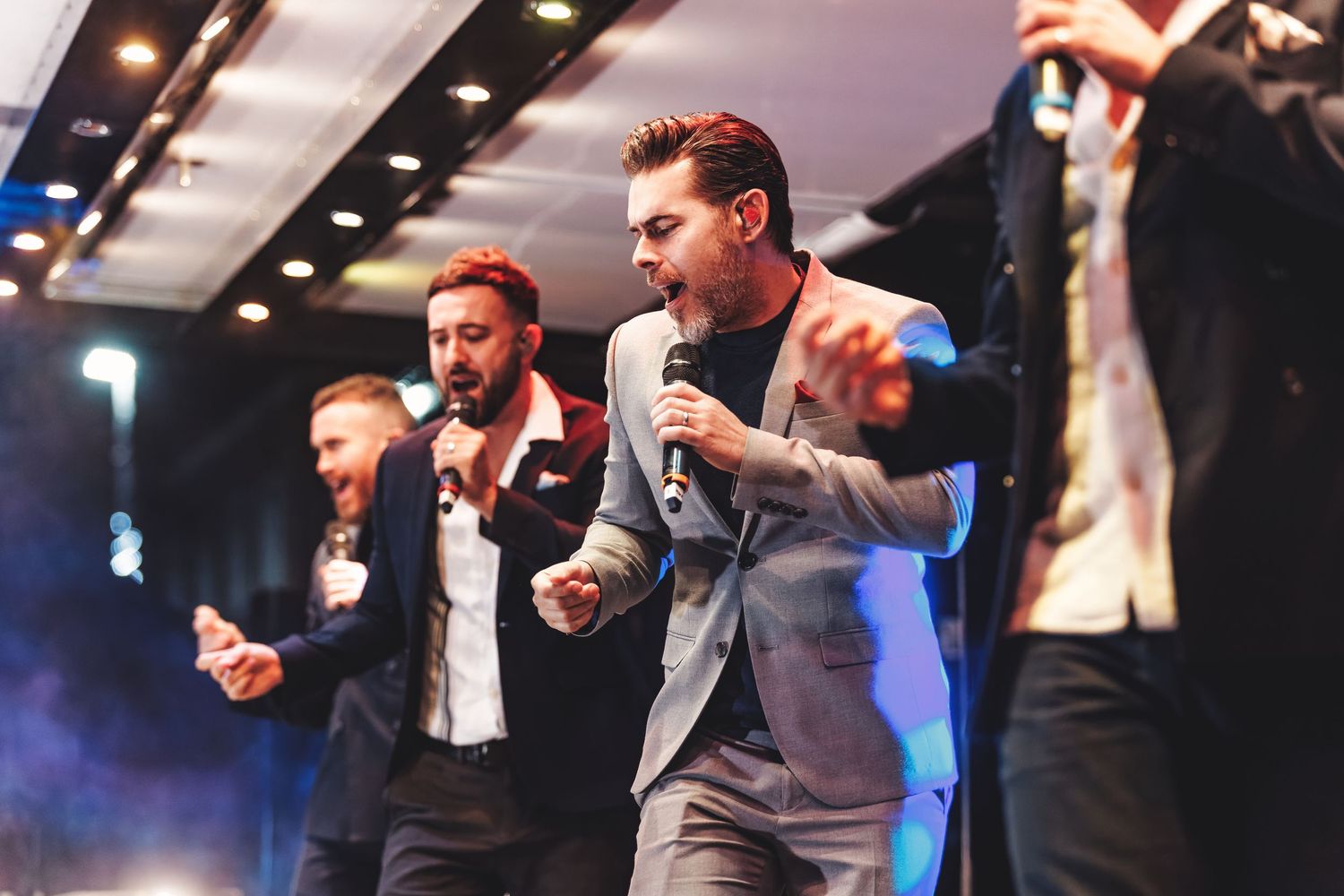 Suited male vocalists performing with microphones on stage at an indoor event with blue and white lighting, live music event photography Southampton