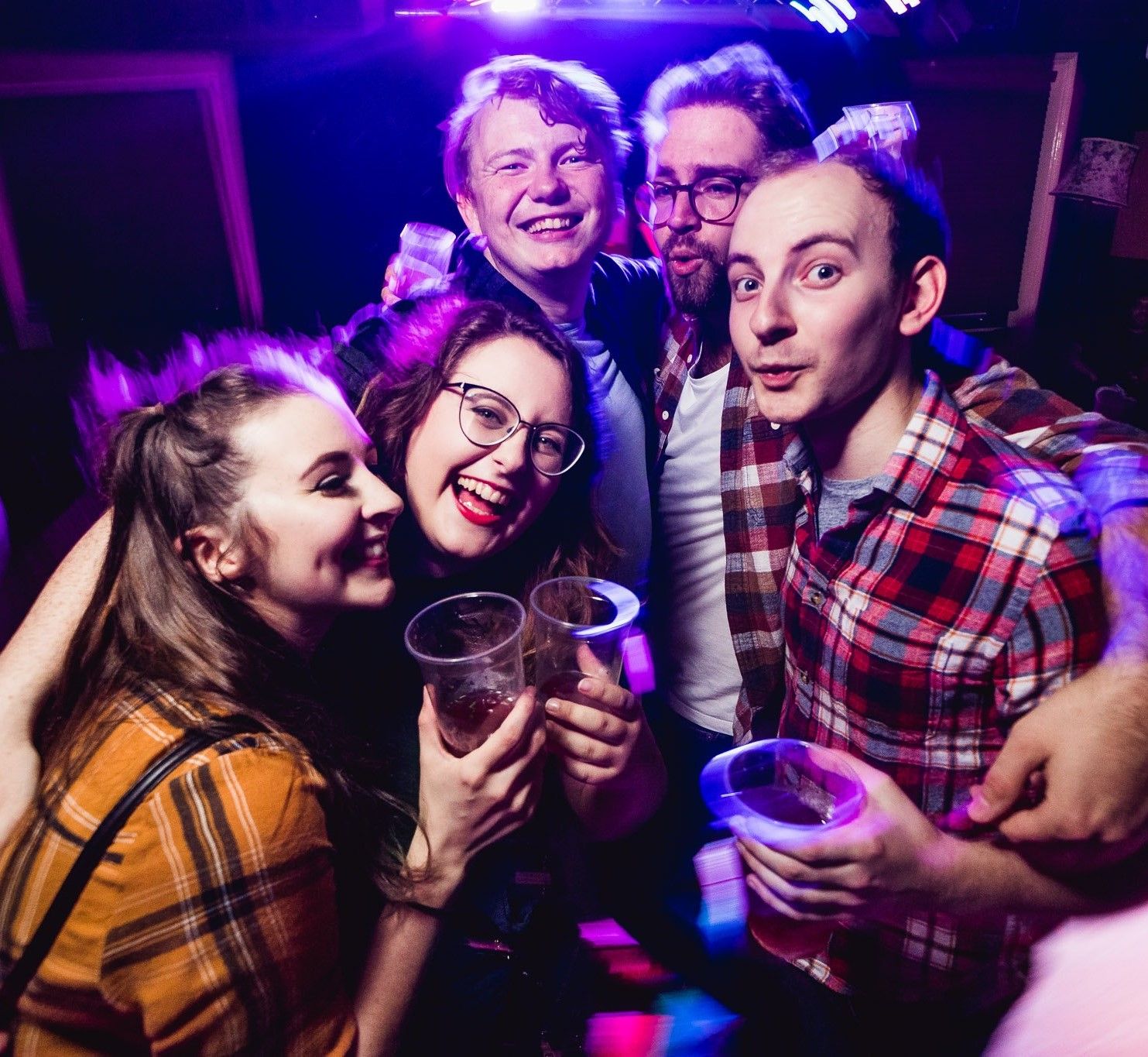 Group of friends with drinks posing at a bar with purple and blue nightclub lighting, nightlife event photography Southampton