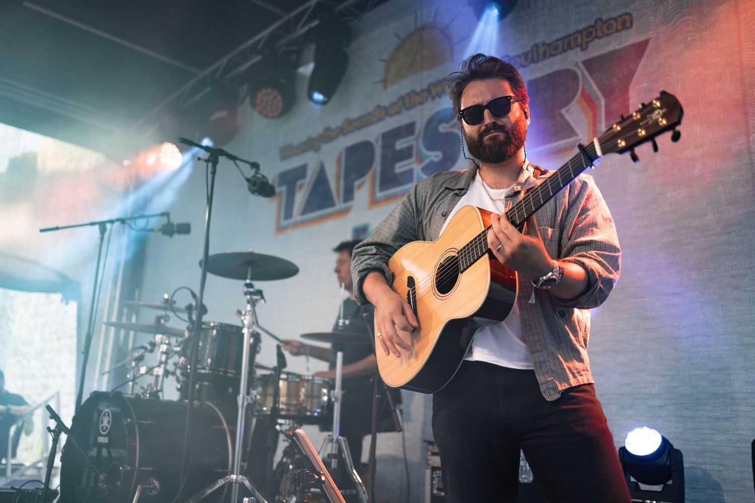 Musician in sunglasses playing acoustic guitar on stage at Tapestry festival Southampton with drummer behind, live music photography