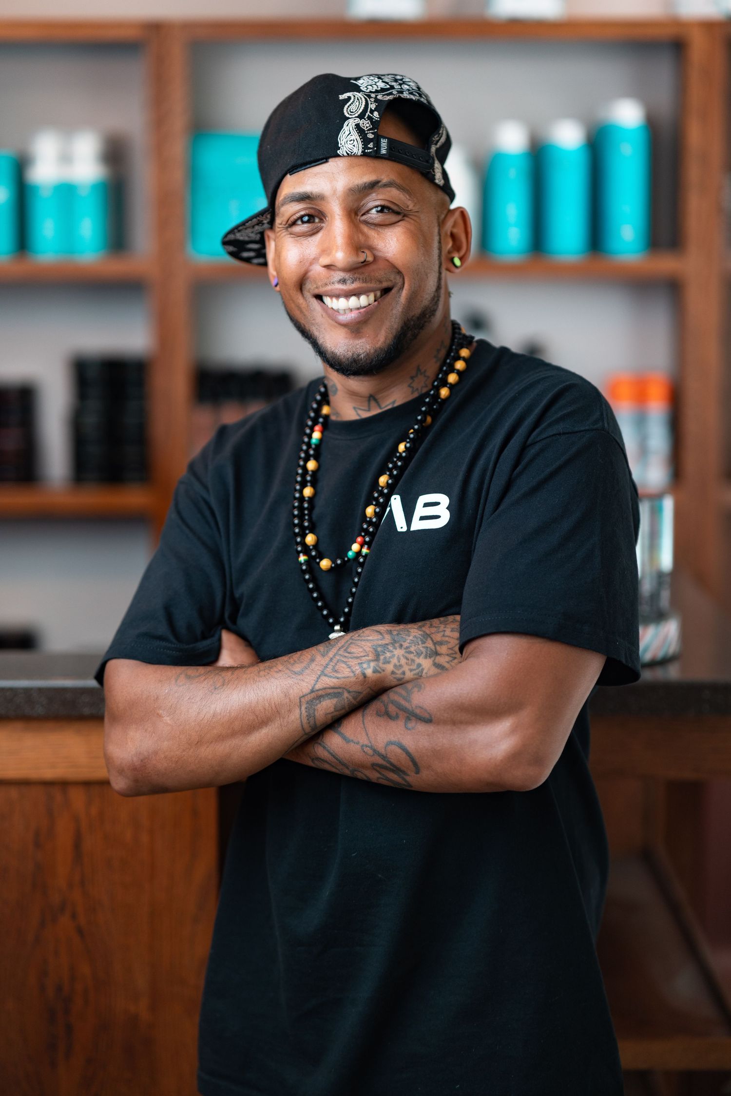 Smiling tattooed barber in a black AB-branded t-shirt, cap and beaded necklace with arms crossed in a salon, team headshot portrait photography Southampton